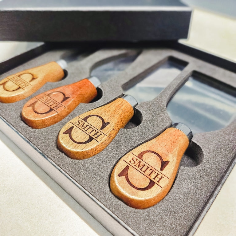 Closeup of set of four cheese knives with wooden handles, engraved with personalized monogram names design, in a black gift box.