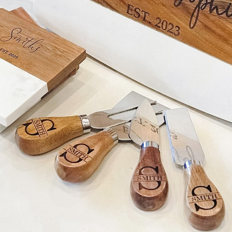 Set of four cheese knives with wooden handles, engraved with personalized monogram names design, placed besides marble coasters and marble serving board
