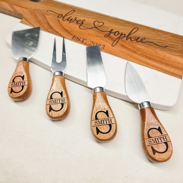 Set of four cheese knives with wooden handles, engraved with personalized monogram names design, placed besides marble serving board