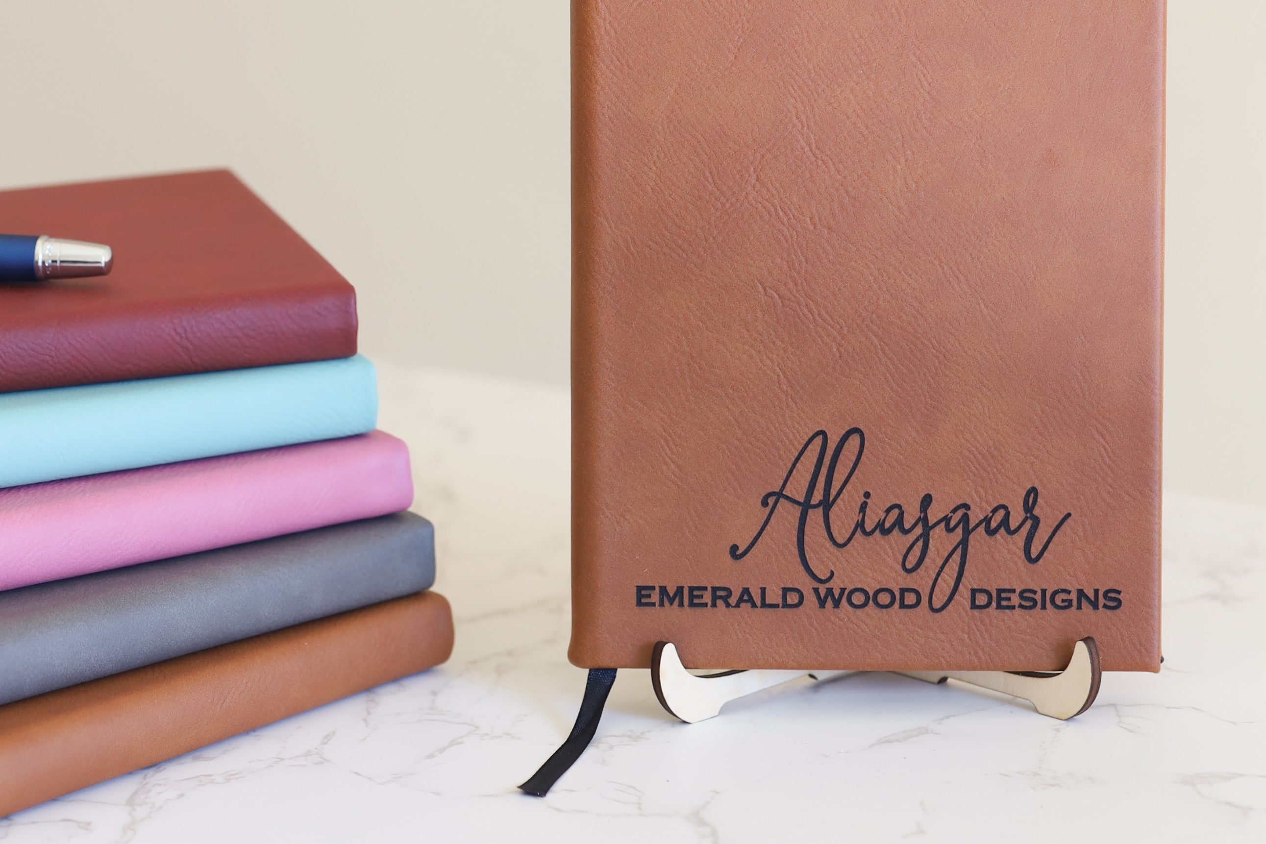 Image of a leatherette journal in rawhide color, with the words "Aliasgar Emerald Wood Designs" laser engraved on it. Besides it is a stack of leatherette journals in various colors of rose, teal, pink, gray, rawhide, with a pen placed on top of the stack.