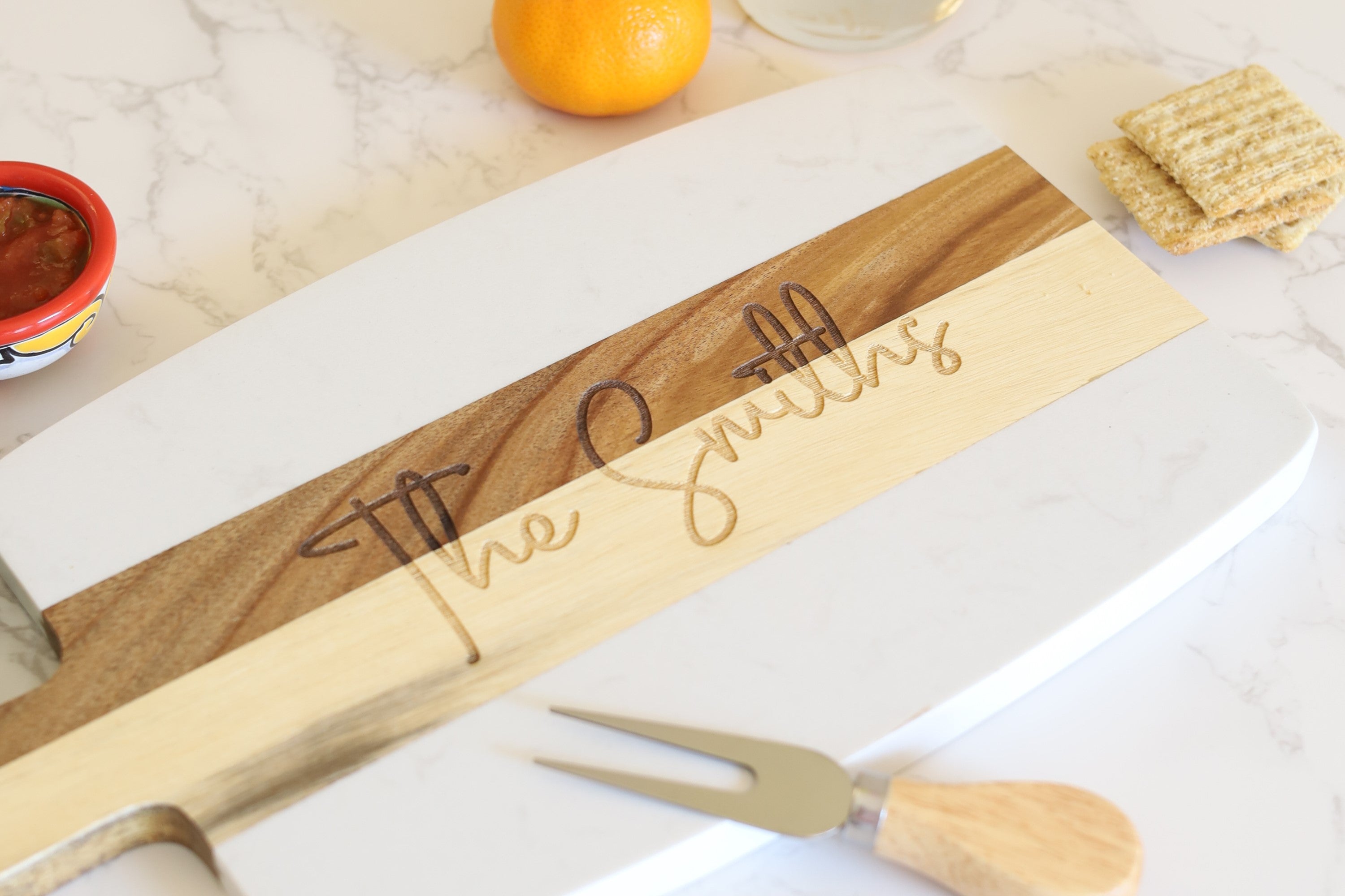 Marble and wood serving board in the shape of a paddle, with the words The Smith laser engraved on the wood part of the board. Image contains accessories of cheese knife, crackers, cheese, juice, fruits, etc.