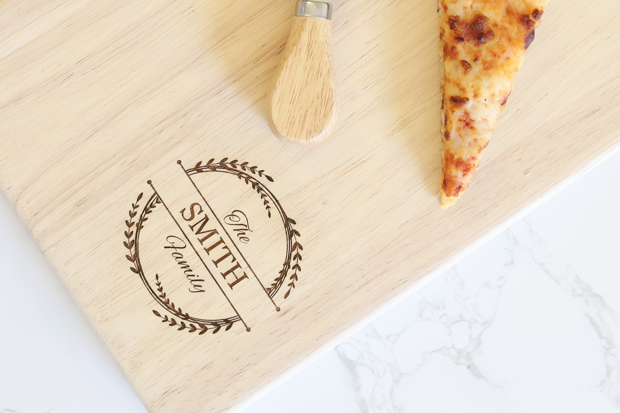 Wooden paddle charcuterie board, with the words "The Smith Family" in artistic font and a wreath design around it, laser engraved on the board. There is also a pizza slice and a cheese knife on the board.