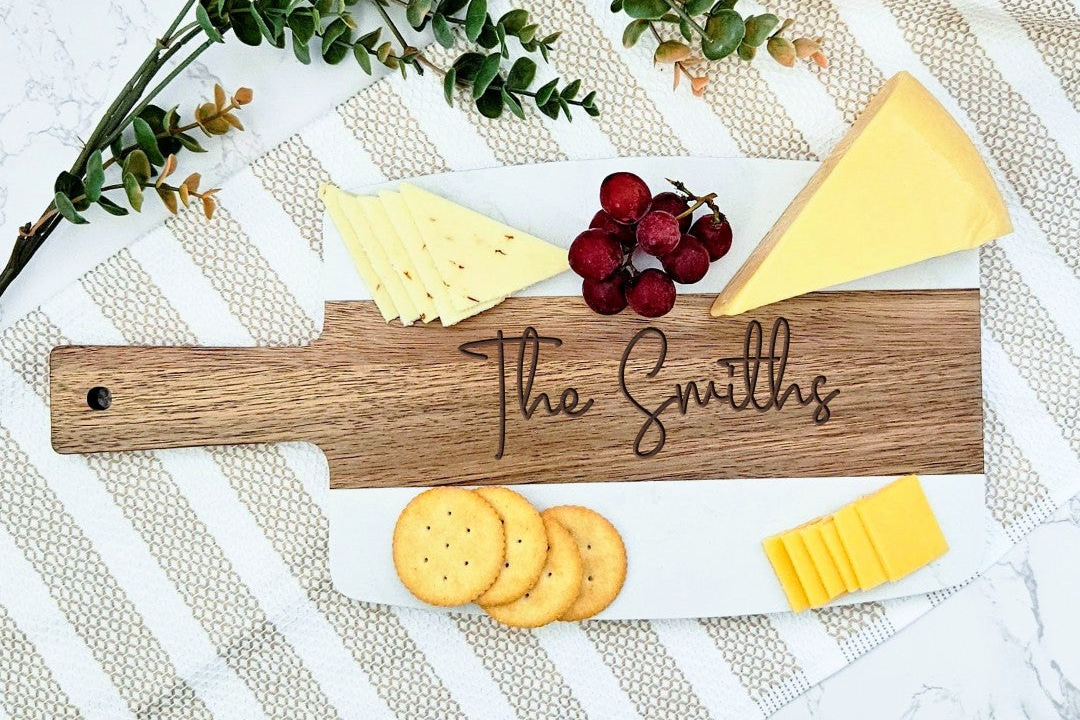 Wooden cheese board with 'The Smiths' engraving, cheese, crackers, and grapes on a marble surface.