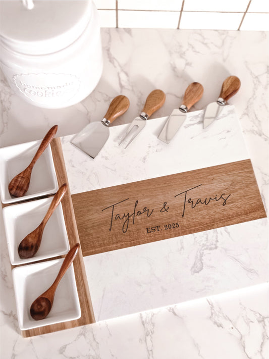 White marble and acacia wood cheese board with engraved with personalized names and dates on a kitchen counter with four cheese knives, three bowls and three spoons.