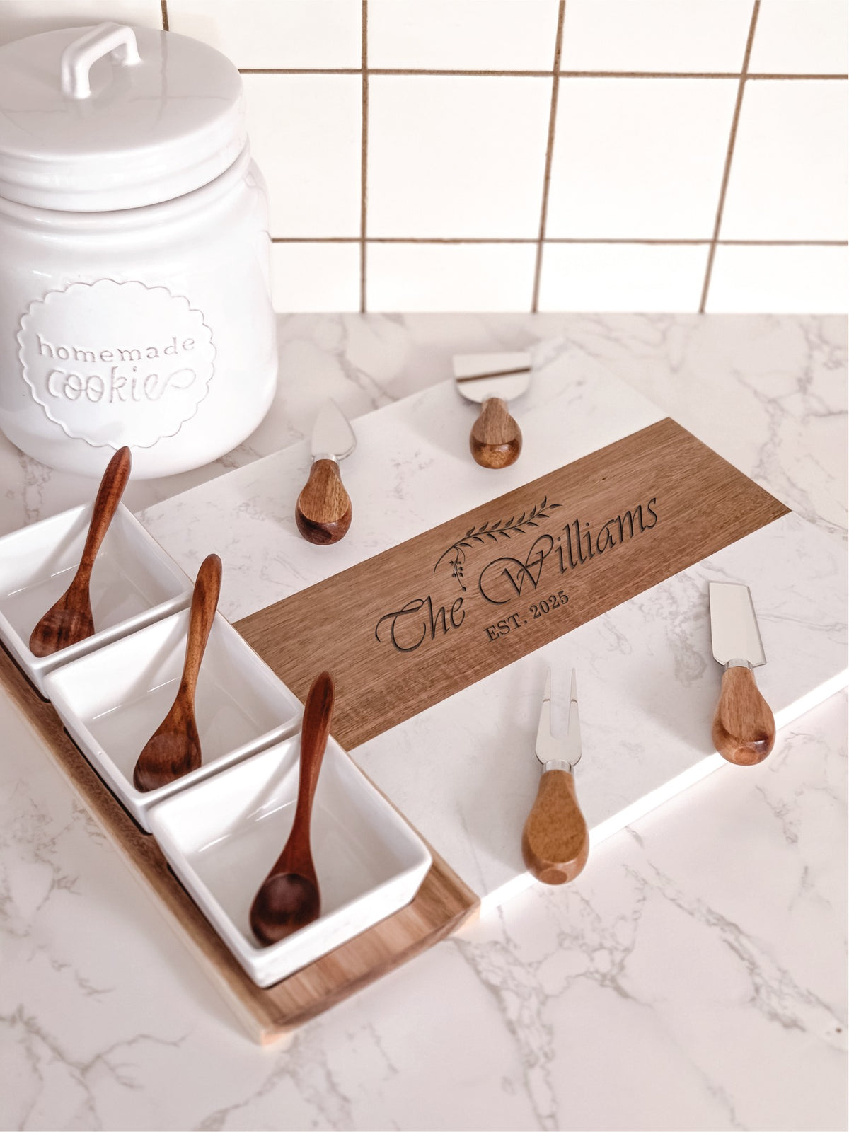 White marble and acacia wood cheese board with engraved with personalized names and dates on a kitchen counter with four cheese knives, three bowls and three spoons.