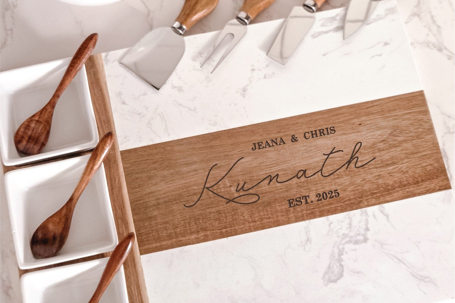 White marble and acacia wood cheese board with engraved with personalized names and dates on a kitchen counter with four cheese knives, three bowls and three spoons.