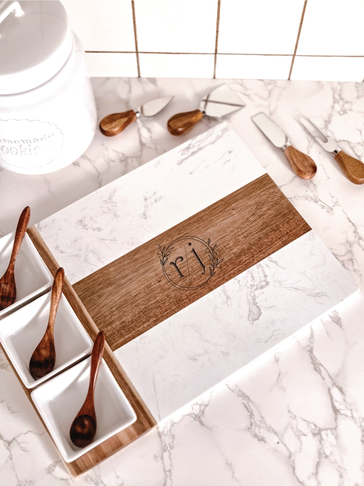 White marble and acacia wood cheese board with engraved with personalized names and dates on a kitchen counter with four cheese knives, three bowls and three spoons.