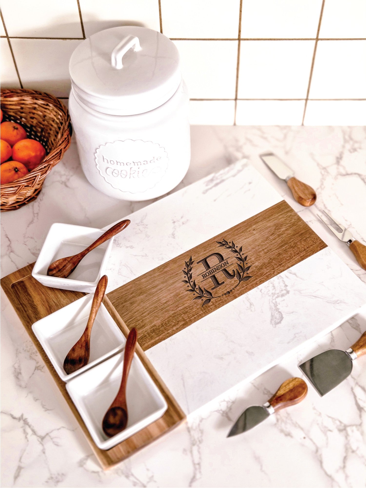 White marble and acacia wood cheese board with engraved with personalized names and dates on a kitchen counter with four cheese knives, three bowls and three spoons.